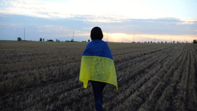 Ukrainian woman walking on barley meadow with a blue-yellow banner on shoulders at sunset. Lady going on wheat field with a flag of Ukraine as symbol of victory against russian aggression. Slowmo