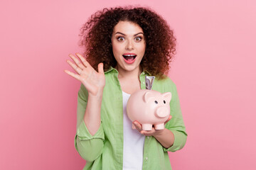 Photo portrait of attractive young woman hold piggy raise hand impressed wear trendy green clothes isolated on pink color background