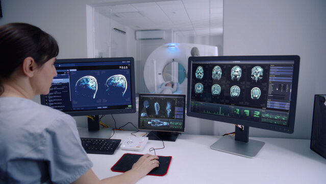 Female radiologist sits in control room and watches at monitors with displayed brain scans results. Professional doctor controls MRI or CT or PET scan with patient undergoing procedure at background.