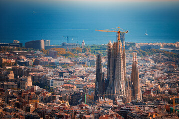 Panoramic view of Barcelona featuring the unfinished Sagrada Familia with its spires and cranes, surrounded by the city's warmhued buildings and the Mediterranean Sea in the backdrop.