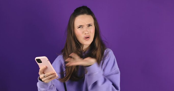 Mad and shock young brunette beautiful woman using mobile phone isolated on purple background. Yelling unhappy woman hold mobile phone. Disappointed sad upset lady horrified impressed news. WTF. Oh no