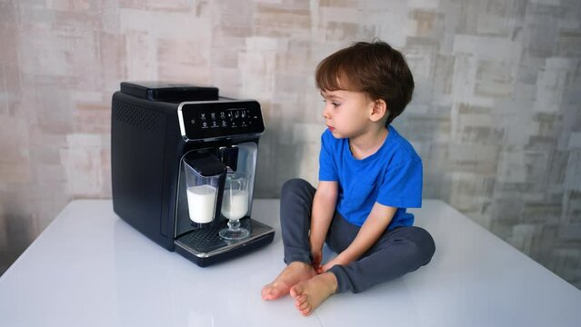 Beautiful Caucasian baby in blue t-shirt sits on the table near the coffee machine. Kid watches milk pouring into the cup from device.