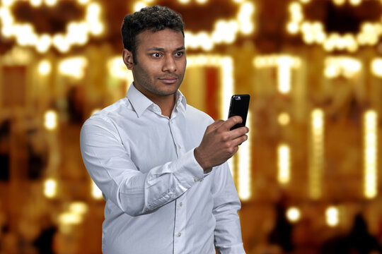Young handsome man wearing white shirt taking picture with mobile phone. Festive bokeh lights in the background.