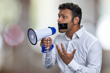 Young man activist with black tape over mouth can not speak into megaphone. Abstract bokeh...