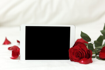 Tablet with black screen mockup for modern electronic invitation or greeting card, red rose flower and petals on white background. St. Valentines day celebration. Women's day. Copy space for text.