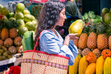 Close-up shot of Latin girl in the middle of a fresh fruit market, with a basket holding a papaya...