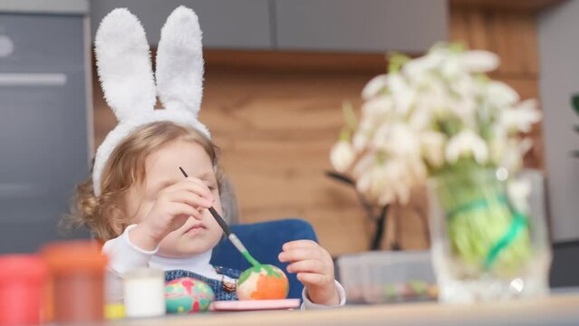 A little girl wearing bunny ears is painting easter eggs in a kitchen