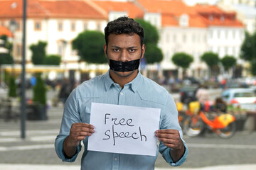 Young indian man with taped mouth holding paper card with handwritten inscription Free speech. Blur city street in the background.