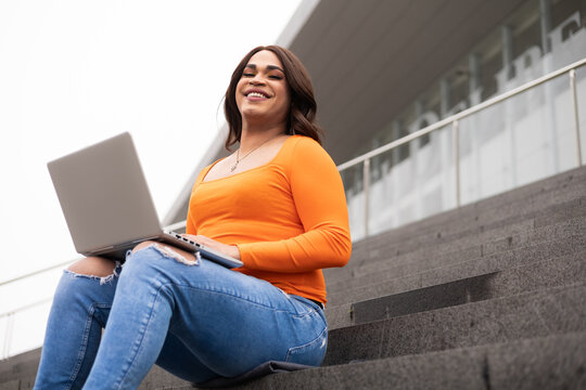 Trans woman student looking to the camera and using laptop in university stairs.
