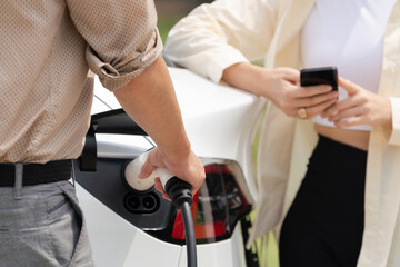 Obraz na płótnie Canvas Young couple use smartphone to pay for electricity at public EV car charging station green city park. Modern environmental and sustainable urban lifestyle with EV vehicle. Expedient