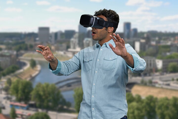 Young shocked man wearing vr glasses outdoor. Blur city skyscrapers in the background. Scared surprised man wearing vr headset.