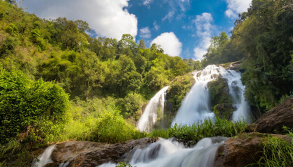 Waterfall flowing through green forest, clear blue sky with fluffy clouds; beautiful nature background
