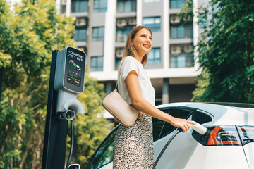 Young woman travel with EV electric car charging in green sustainable city outdoor garden in...
