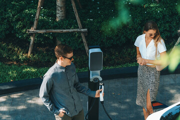 Young couple travel with EV electric car charging in green sustainable city outdoor garden in...