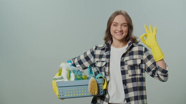A Portrait of a Smiling Young Woman in Yellow Protective Rubber Gloves Holding a Box With Cleaning Products and Shows Okay Sign on the Gray Background. Pet-friendly Cleaning Solutions.