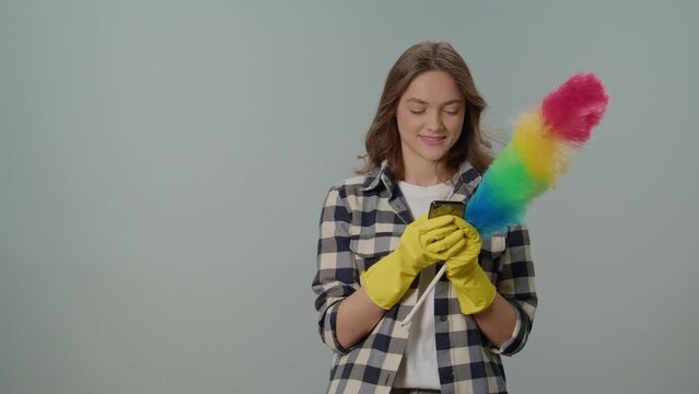 A Happy Young Woman in Yellow Gloves and With a Dustpan, Rejoices at Success on the Phone on the Gray Background.Wireless and App-controlled Cleaning Appliances.