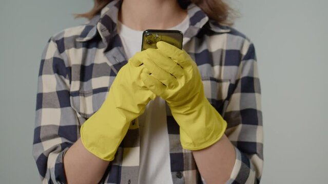 Close-up of the Female Hands in Yellow Gloves with a Smartphone. A Young Woman Housewife in Yellow Gloves Texting With Someone on Her Smartphone ,on the Gray Background.Virtual Cleaning Consultations.