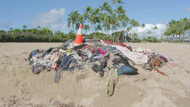 A stack of dirty sandy shore environmental pollution ecological problem, clothes and a traffic cone on a sandy beach with scenic views of the sky clouds and palm trees plants in Oahu Hawaii 