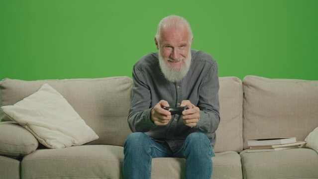 Green Screen. An Old Man With a Gray Beard Plays Computer Games With a Joystick. An Elderly Man Plays PlayStation and Wins. Tech Nostalgia and Generational Divide.