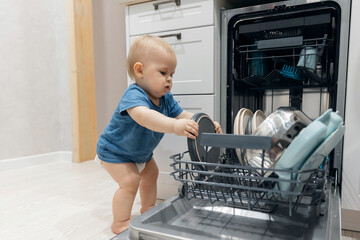 Little child helping to unload dish in dishwasher. Concept Independent baby boy working in kitchen