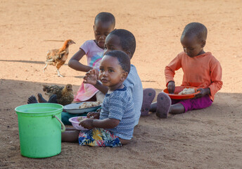 hungry village african children in the yard , sited on the ground eating staple food from plastic plates - obrazy, fototapety, plakaty