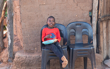 hungry village african child in the yard , sited on a chair eating staple food from a plastic...