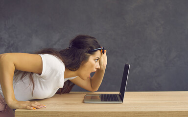 Young caucasian shocked emotional woman with surprised face looking at laptop screen. Side view of a woman sitting at a table leaning closer to a laptop screen to make sure she really saw it. - obrazy, fototapety, plakaty