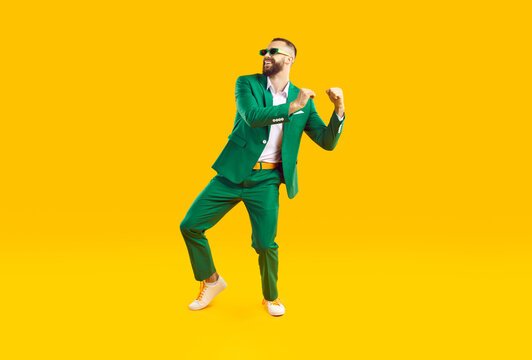 Cheerful and stylish young man is having fun dancing at St. Patrick's Day party. Cool Caucasian man in fashionable green suit and sunglasses smiling while dancing on orange background. Full length.