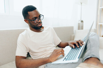 Young African American freelancer sitting on a modern sofa in his home office, happily working on...