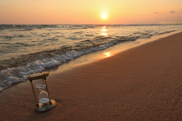An hourglass (sand clock) lies on the beach sand with the background of the sea and the setting sun...