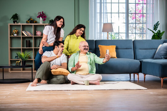 Happy smiling indian senior parents with adult son using smartphone while sitting on sofa at home.