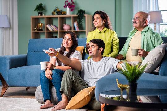 Indian family of four sitting on couch watching TV, operating with remote control - Powered by Adobe