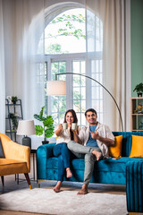 Cheerful young beautiful Indian couple having coffee together on sofa at home..