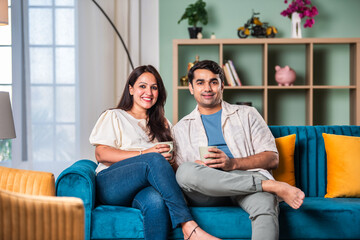 Cheerful young beautiful Indian couple having coffee together on sofa at home..