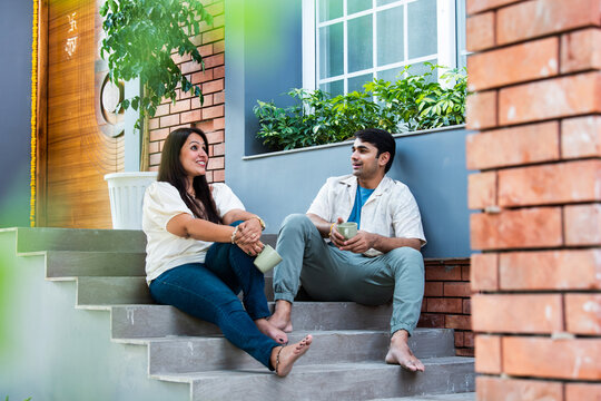Cheerful young beautiful Indian couple having coffee together on stairs at home..