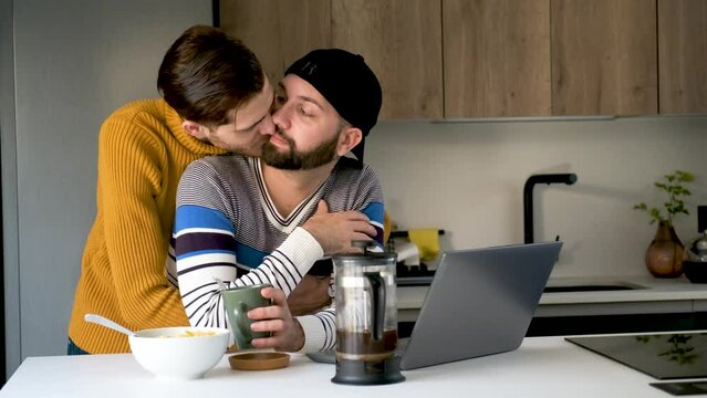 Man working on a laptop and his boyfriend hug and kiss him.