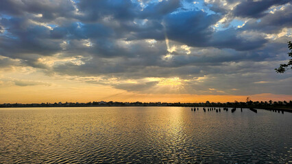 Obraz premium Sunrise casts a soft golden light piercing through clouds above the still waters of a serene lake at dawn