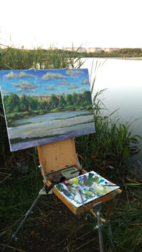 Easel sketchbook with palette oil painting on background of a river water pond plain air. The artist workplace in open air.