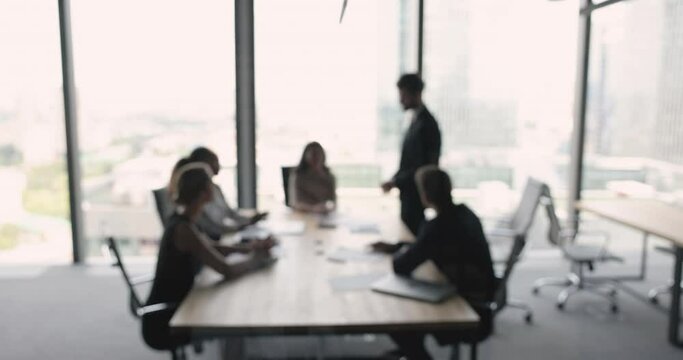 Business team meeting at large table in office space with large window, talking, brainstorming on ideas, listening to standing speaker. Negotiations of partners, teamwork blurred background