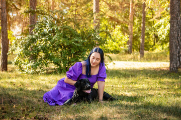 Naklejka na ściany i meble A young woman in a purple dress sits next to a black Labrador retriever against the backdrop of the park.