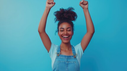 Young woman celebrates success after winning prize. Full body length happy joyful beautiful slim African American girl in casual wear standing on bright blue background, raising hands up and cheering - obrazy, fototapety, plakaty