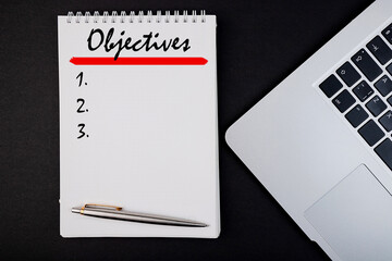 Objectives word concept written in a notebook with a pen and laptop, top view.
