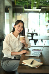 Fototapeta premium Smiling millennial female worker holding paper cup and working with laptop at coffee shop