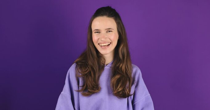 Friendly looking positive straight hair woman shows shaka sign gestures indoor being in high spirit, wears fashionable purple hoody isolated on violet background. Girl show hi or hello gesture. Jumbo.