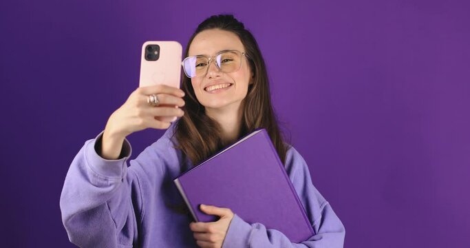 Happy brunette woman in glasses smiling and holding purple book while taking selfie photo isolated over violet background. Blogger woman student make photo or video.