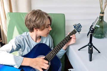 Young teenage boy streaming video online for social media blog or webinar, while playing guitar, making online music lesson, teenagers in cyberspace concept