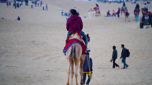 slow motion shot of young boy leading camel with tourists sitting on it while people roam around at sunset in thar desert in Sam Jaisalmer Rajasthan