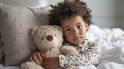 A young child with curly hair wearing a patterned onesie cuddling a light brown teddy bear resting on a bed with a geometric patterned pillow.