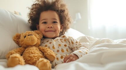Fototapeta premium A joyful child with curly hair wearing a white onesie with yellow patterns sitting on a bed with a brown teddy bear.