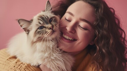 A woman with long hair wearing a yellow sweater smiling and holding a fluffy white cat with a gray face against a pink background.